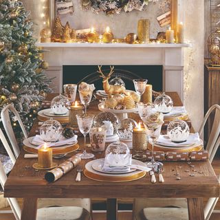 A dining table set for dinner with metallic golden decor also seen on the mantelpiece and Christmas tree in the background