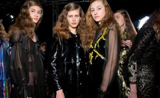 Three women, one in a sheer black dress, one in a black shiny coat, and one in a black velvet dress with sheer sleeves and yellow ruffle designs