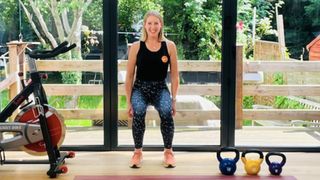 Fitness trainer Maddy Biddulph faces the camera, smiling, as she performs a wall sit. Her back is against a full-length window, her knees are bent at 90 degrees and her arms hang by her side. In front of her, we can see an exercise mat, three kettlebells and an exercise bike. Behind her is a leafy garden.