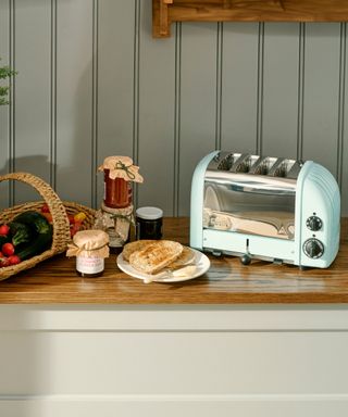 A light blue Dualit toaster on a kitchen counter with jam, toast, and a basket next to it, with gray wall paneling behind it
