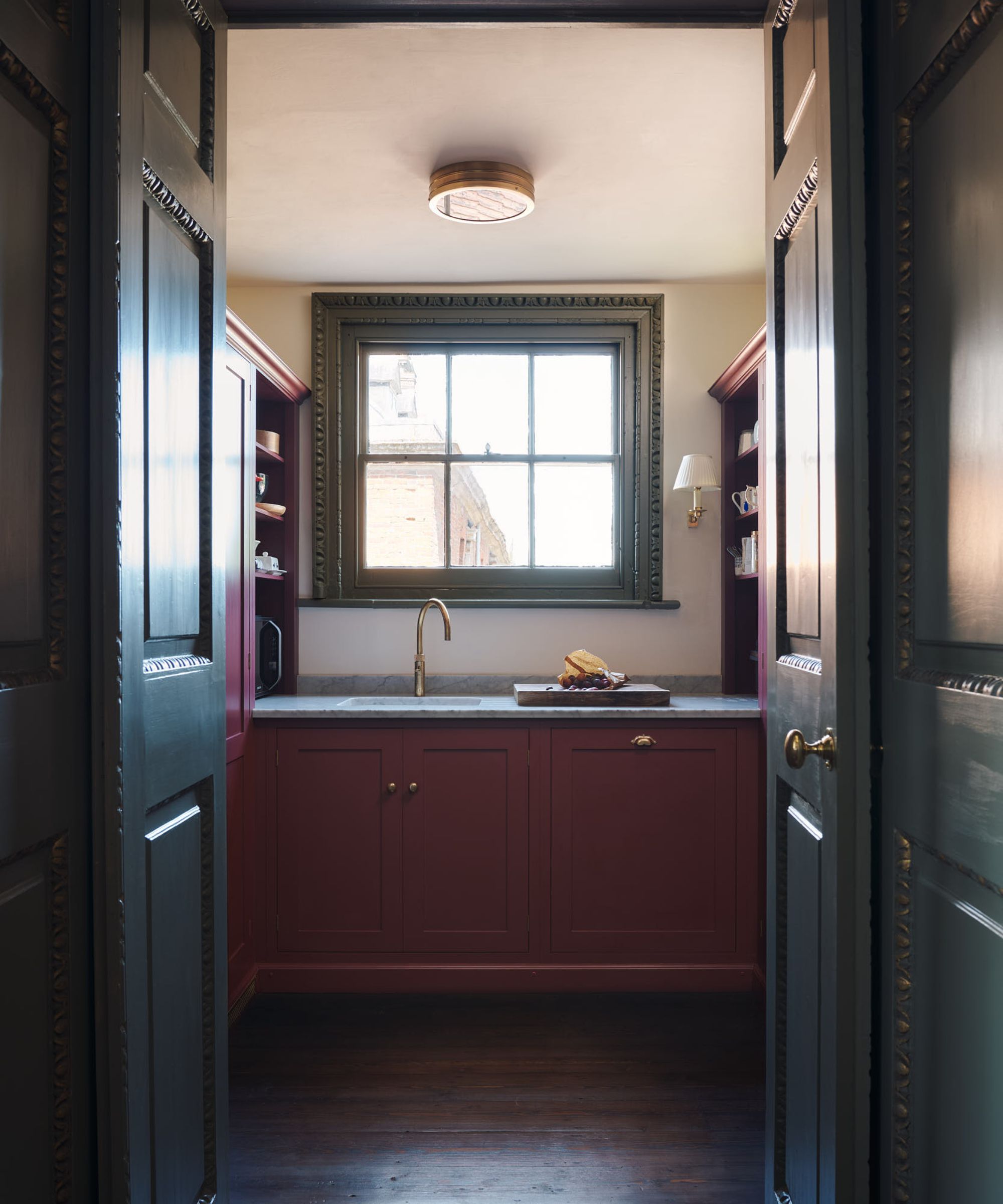 A stately home pantry with red cabinets, white walls, and a green window frame