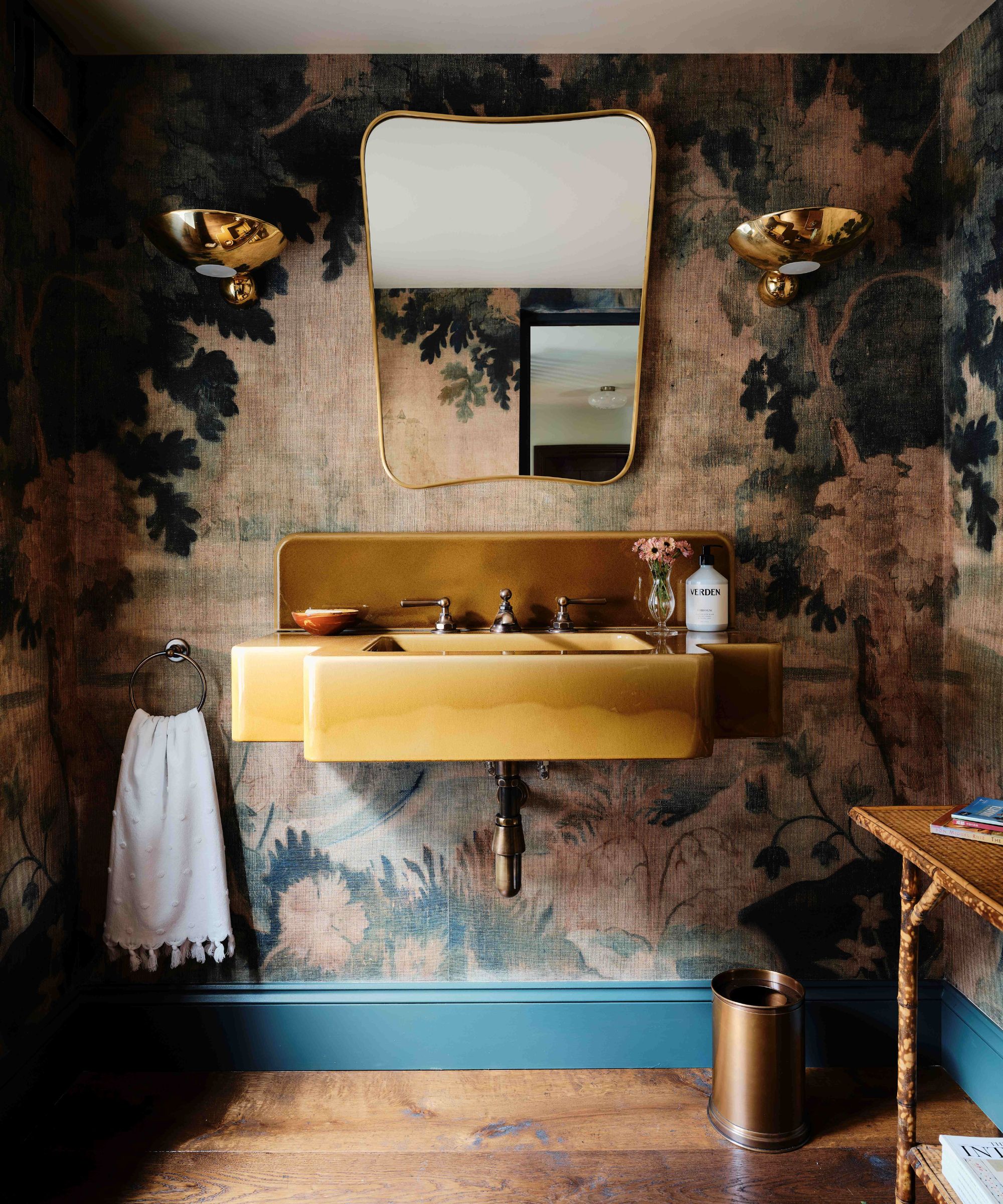 powder bathroom with an earthy moody botanical mural wallpaper, blue painted baseboards, an ochre floating sink, mid century mirror and brass sconces