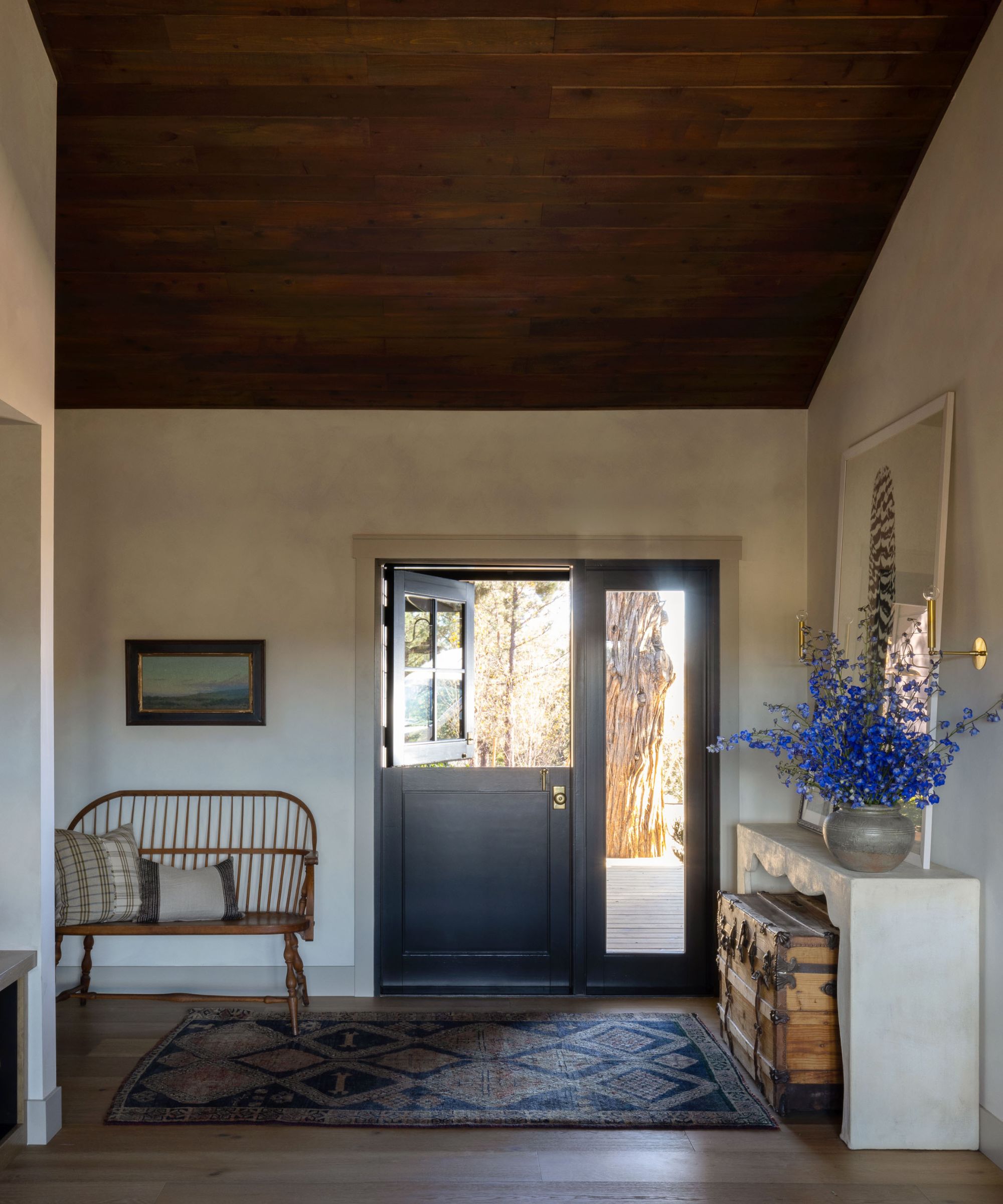An entryway with a black Dutch door, vintage rug, and wooden bench with plaid cushions. There is a console table with a chest underneath.