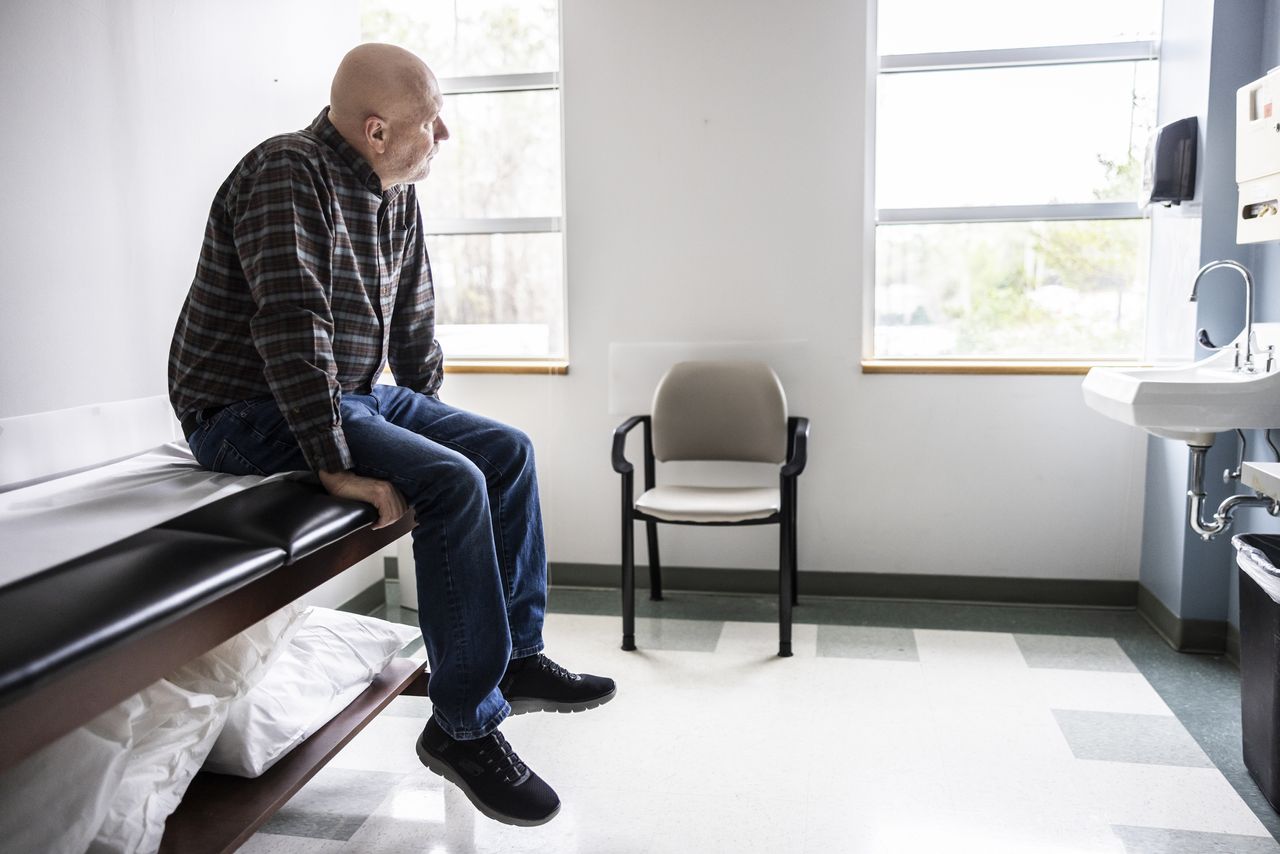 An older man sits on a doctor&#039;s office seat waiting to be seen.