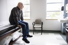 An older man sits on a doctor's office seat waiting to be seen.