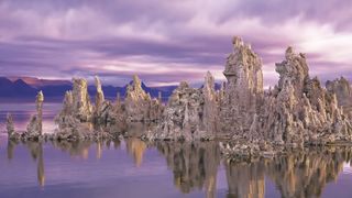 strange white rock formations reflected in water below. The dawn sky is light pink and purple.