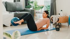 A woman exercises at home an a mat. She is on her back, propped up on her elbows, legs elevated, knees bent and a small Pilates ball held between her feet. There is a kettlebell and water bottle near the mat and in the background we see a couch and leafy plant.