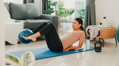 A woman exercises at home an a mat. She is on her back, propped up on her elbows, legs elevated, knees bent and a small Pilates ball held between her feet. There is a kettlebell and water bottle near the mat and in the background we see a couch and leafy plant.