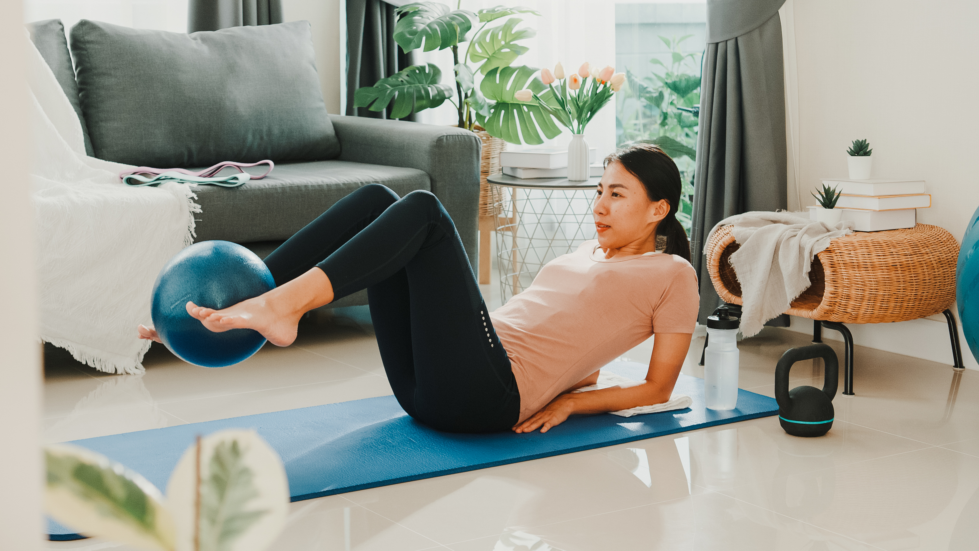 A woman exercises at home an a mat. She is on her back, propped up on her elbows, legs elevated, knees bent and a small Pilates ball held between her feet. There is a kettlebell and water bottle near the mat and in the background we see a couch and leafy plant.
