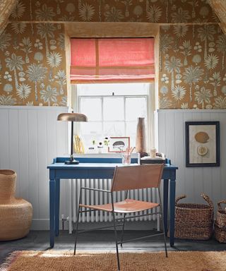 Blue desk and red metal chair in front of window with red Roman blind, floral wallpaper.