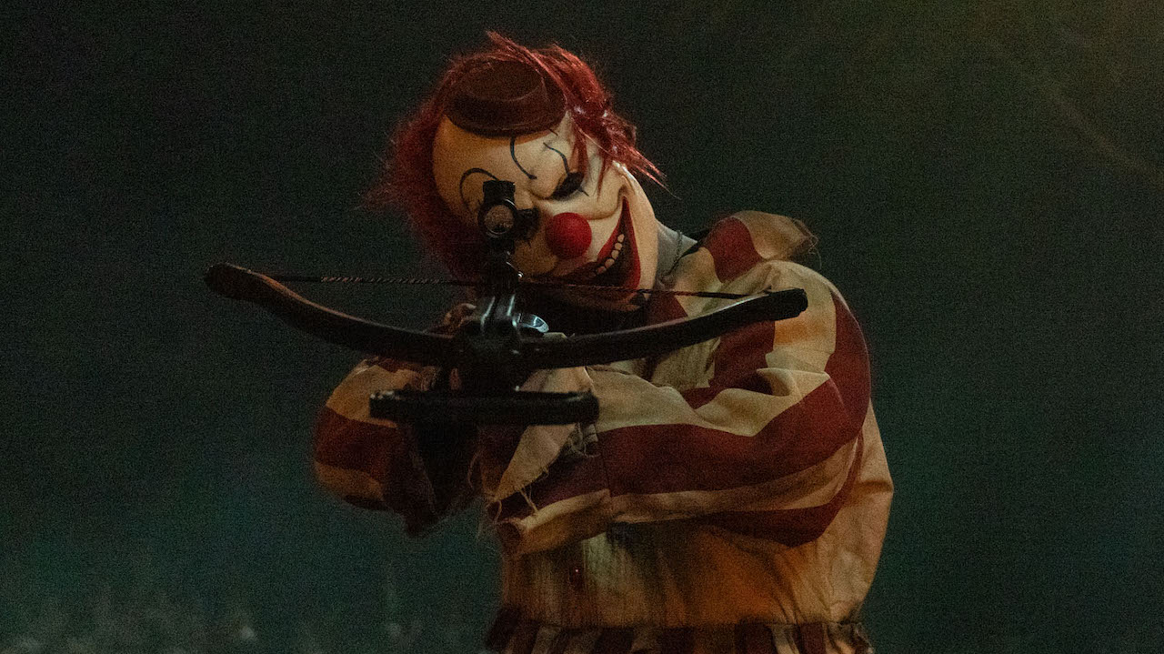 Clown aiming a crossbow toward camera in Clown in a Cornfield