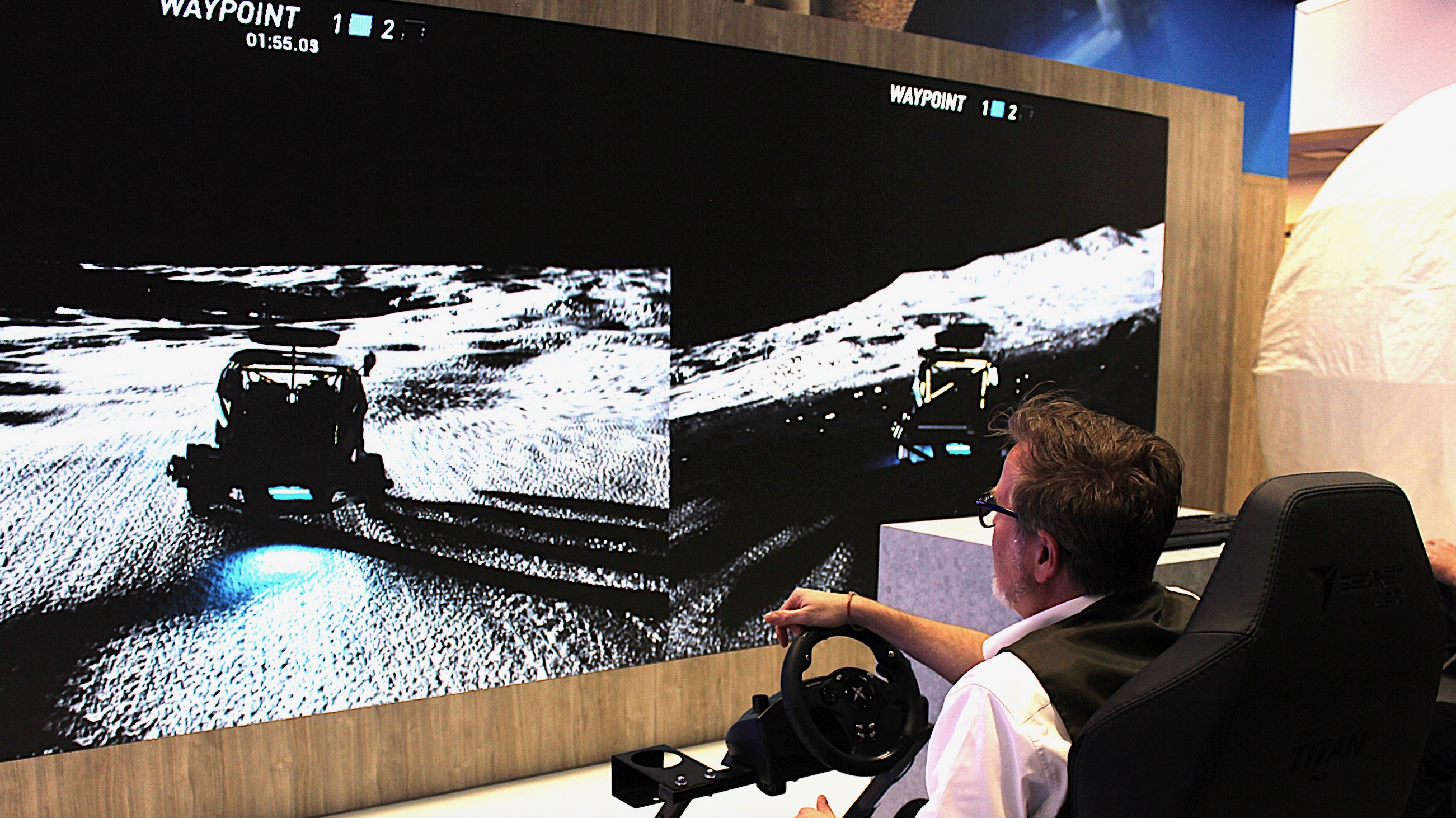 a well-dressed man seated in a chair drives a moon rover in a simulation here on earth