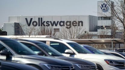 Volkswagen vehicles lined up in a parking lot with company logo and branding pictured on side of an assembly plant in background.