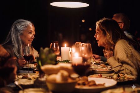 Two older women friends smile and lean towards each other as they talk at a dinner party.