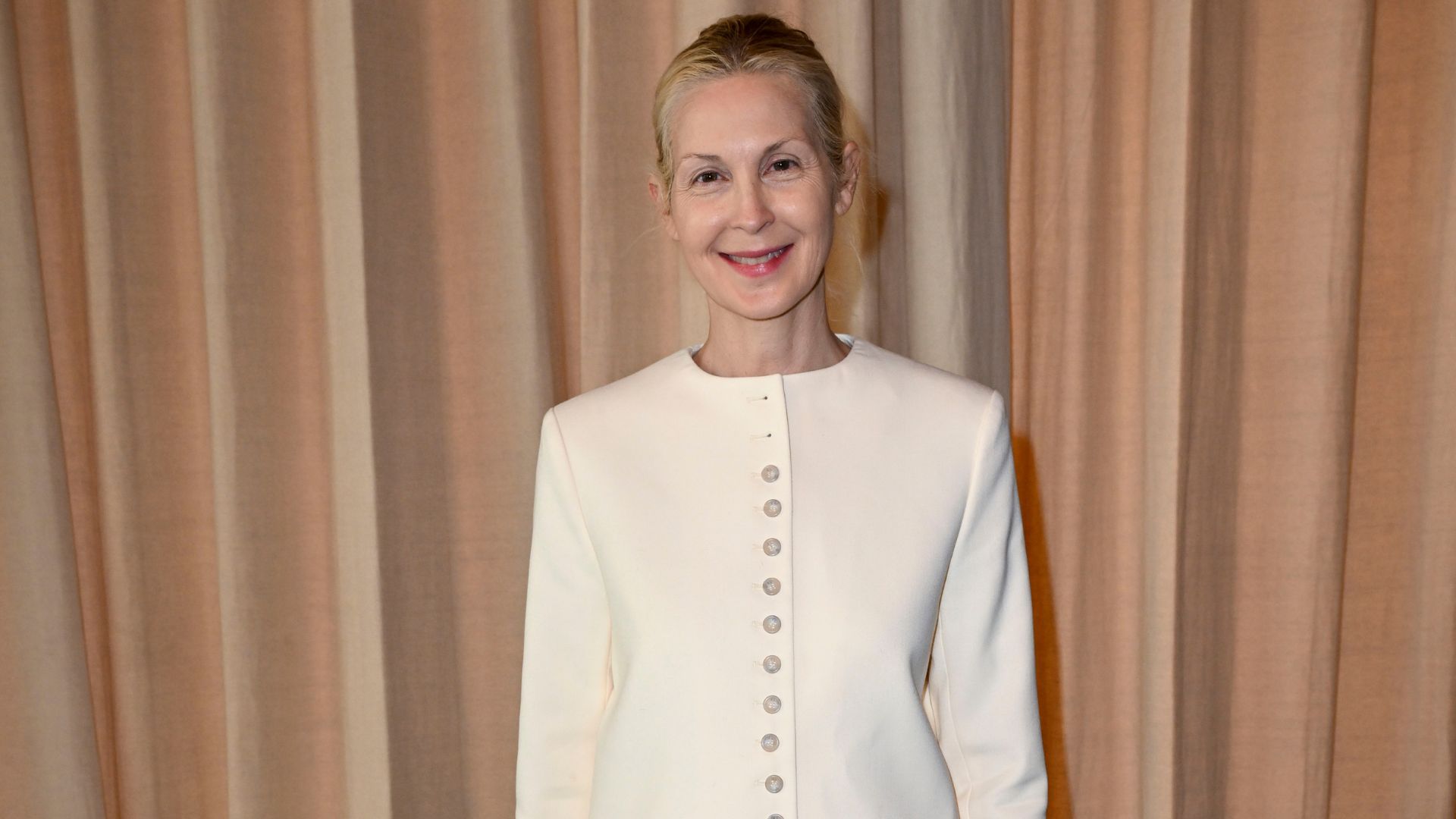 Kelly Rutherford poses for a photo wearing white trousers with beige heels and a white jacket.