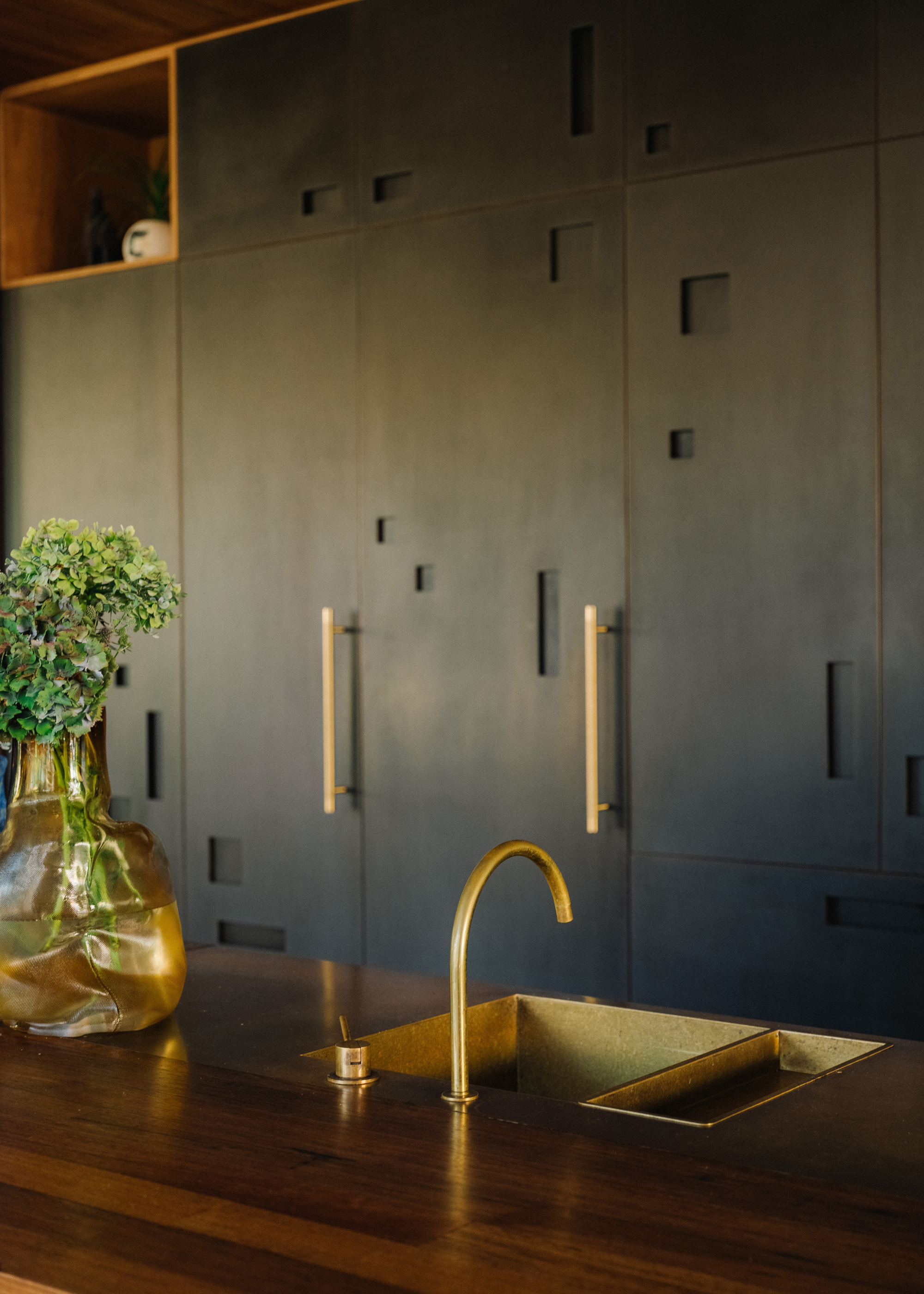 A kitchen with a wood island featuring a brass sink, a vase of flowers, and dark cabinetry