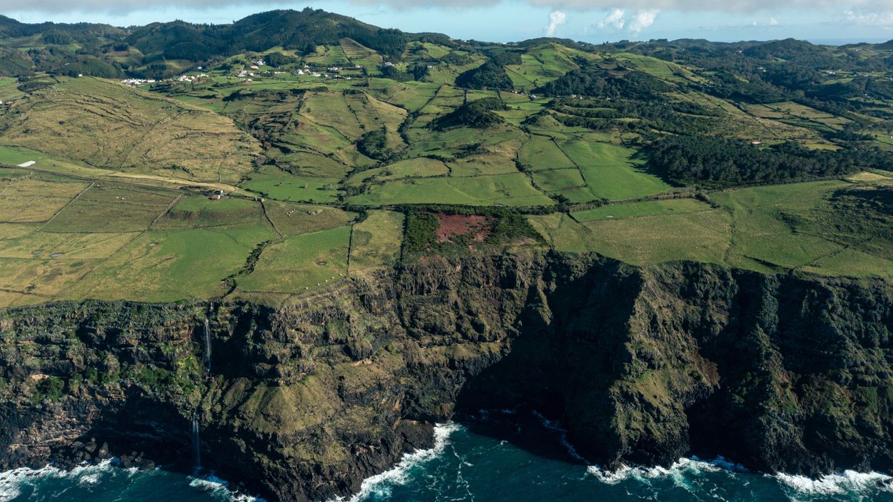 a blue ocean crashes against the bottom of a high cliff that gives way to green fields stretching to mountains in the distance.