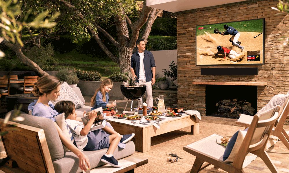 A family on a patio outside. The mother reads books with a toddler, while a little girl plays at a table. The father is watching the baseball game on a Samsung TV while cooking at a grill.