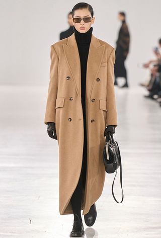 One of the best jacket coat brands, Max Mara, is shown in an image of a model wearing a long camel coat with a black turtleneck and black leggings and black loafers