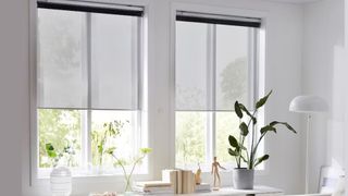 En ljus bild på ett vardagsrumsfönster med växter och böcker på en sideboard framför, med smarta persienner från IKEA som täcker fönstren.