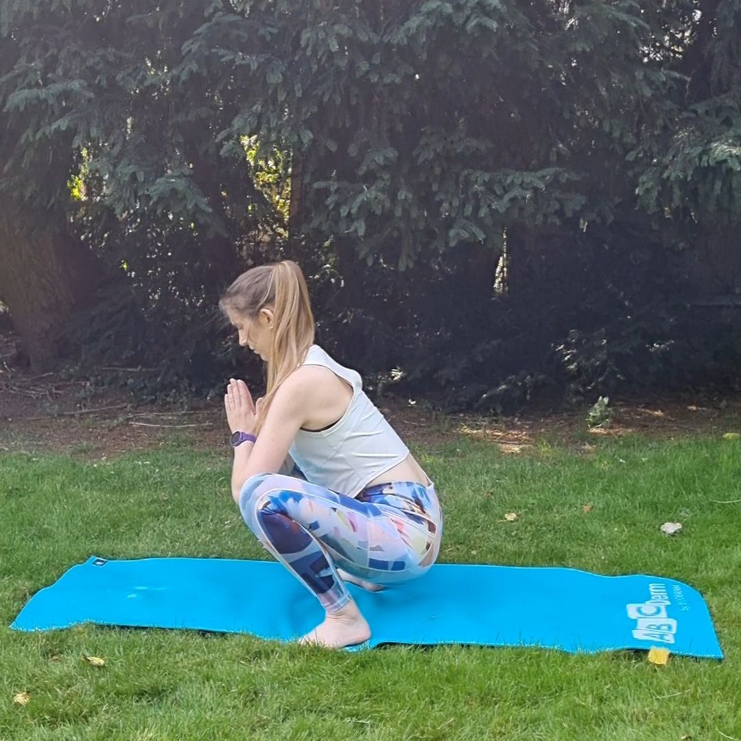 Fitness editor Ruth Gaukrodger performs a deep squat to forward fold. She is in sportsgear and exercising on a yoga mat outdoors. In this picture, she is performing a deep squat, with her knees and hips bent and her hands in a prayer position in front of her chest.