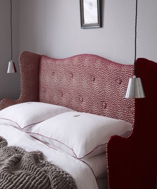 A close up shot of a bed with an embossed red and white headboard with white bedding and two pendant lamps