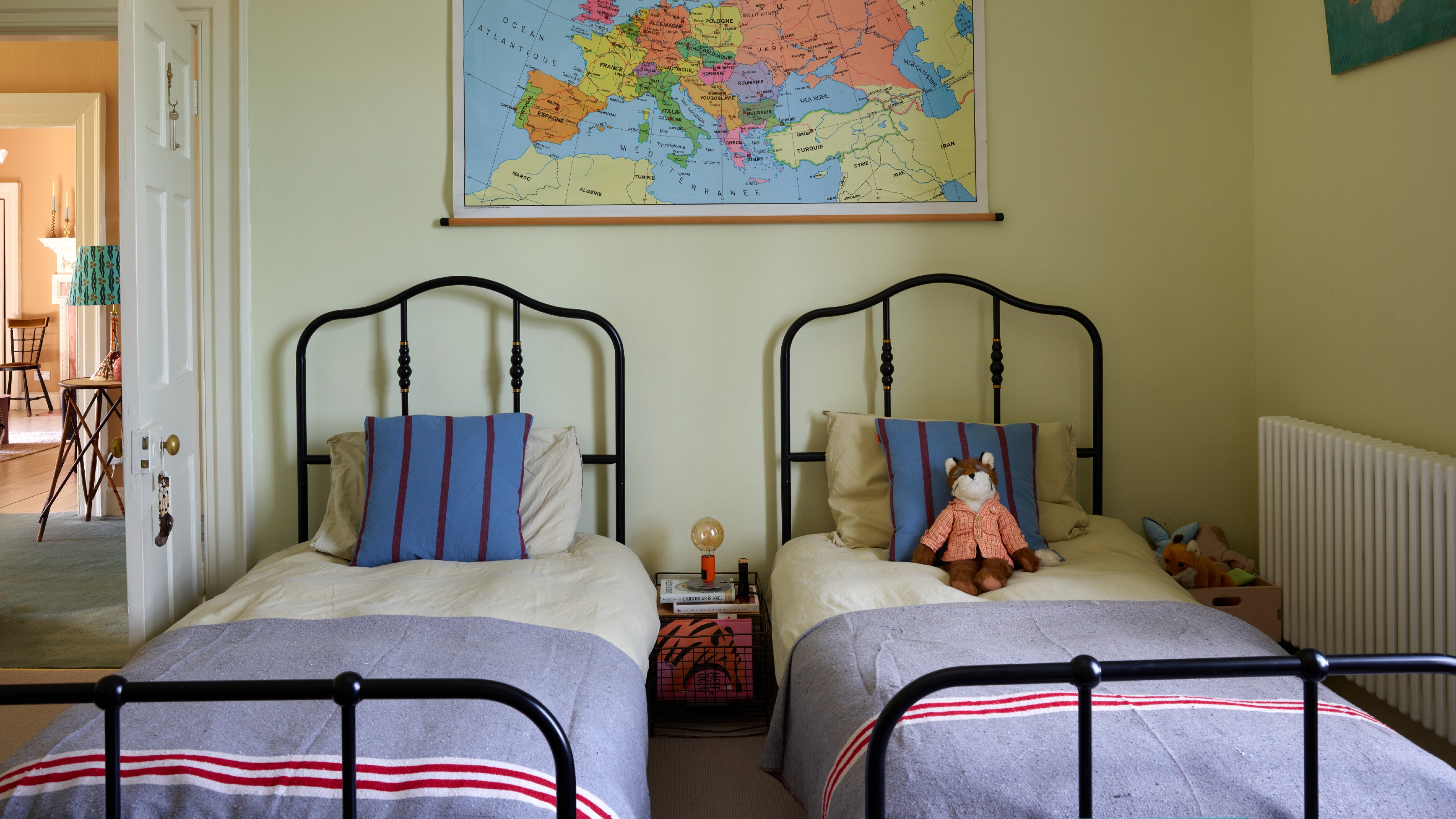 A light yellow-painted shared children's room with two black metal single beds and a map hanging above them