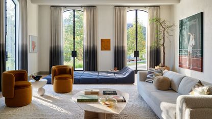 Image of a large living room with tall ceilings and three large glass doors with ombre curtains. There is a boucle sofa and two burnt orange accent chairs. A floor lounger is in the back of the room.