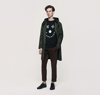 A male model wearing burgundy pants, a dark green jacket and a black shirt with white dots on it.