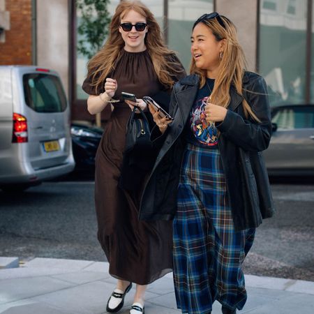 Two women talk and walk in the streets of London while wearing fashionable outfits. 
