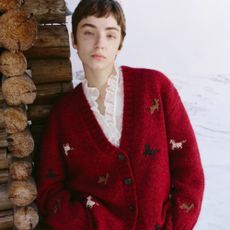 Young woman with short hair wearing cosy red Fabrique cardigan and white ruffle shirt, stood by snow and logs.