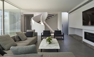 The lounge area of a residential home featuring an L shaped grey sofa with cushions/pillows, center rectangular white table, 2 black arm chairs. a black tv and fire place and a white stair case