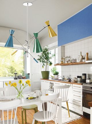 A midcentury modern style white kitchen with traditional white wooden dining set, colorful ceiling lighting with blue, yellow and green shades, blue walls and white square tiles