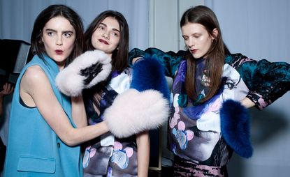 Three female models wearing looks from MSGM's collection. One model is wearing a blue sleeveless jacket and pink and black fluffy mittens. Next to her are two models wearing grey, blue and pink pieces with one model wearing blue fluffy mittens