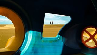 View through colorful playground tunnel framing a couple with a dog on dunes under a blue sky. Bright, playful, and serene atmosphere