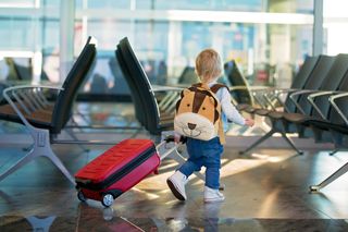 toddler wearing teddy backpack pulling a wheelie suitcase through an airport waiting area