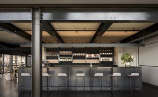 The restaurant's grey coloured bar with a display of wine bottles, 7 white bar stools and a plant filled vase on the right end of the bar.