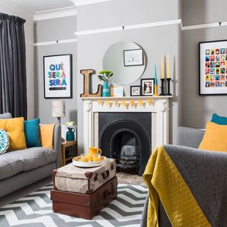 a grey living room with yellow and blue accents such as cushions, throw, candles and yellow bunting, with two suitcases used as a coffee table, on top of a grey and white zig zag rug