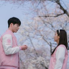 A man and a woman (Seo Kang-jun as Seo Eun-ho and Jisoo as Seo Mi-rae) wear pink and white Letterman jackets while standing outdoors in front of a cherry blossom tree, in 'Boyfriend on Demand.'