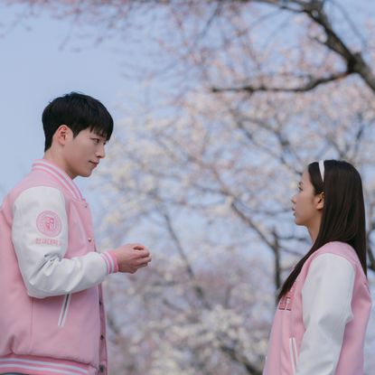 A man and a woman (Seo Kang-jun as Seo Eun-ho and Jisoo as Seo Mi-rae) wear pink and white Letterman jackets while standing outdoors in front of a cherry blossom tree, in 'Boyfriend on Demand.'