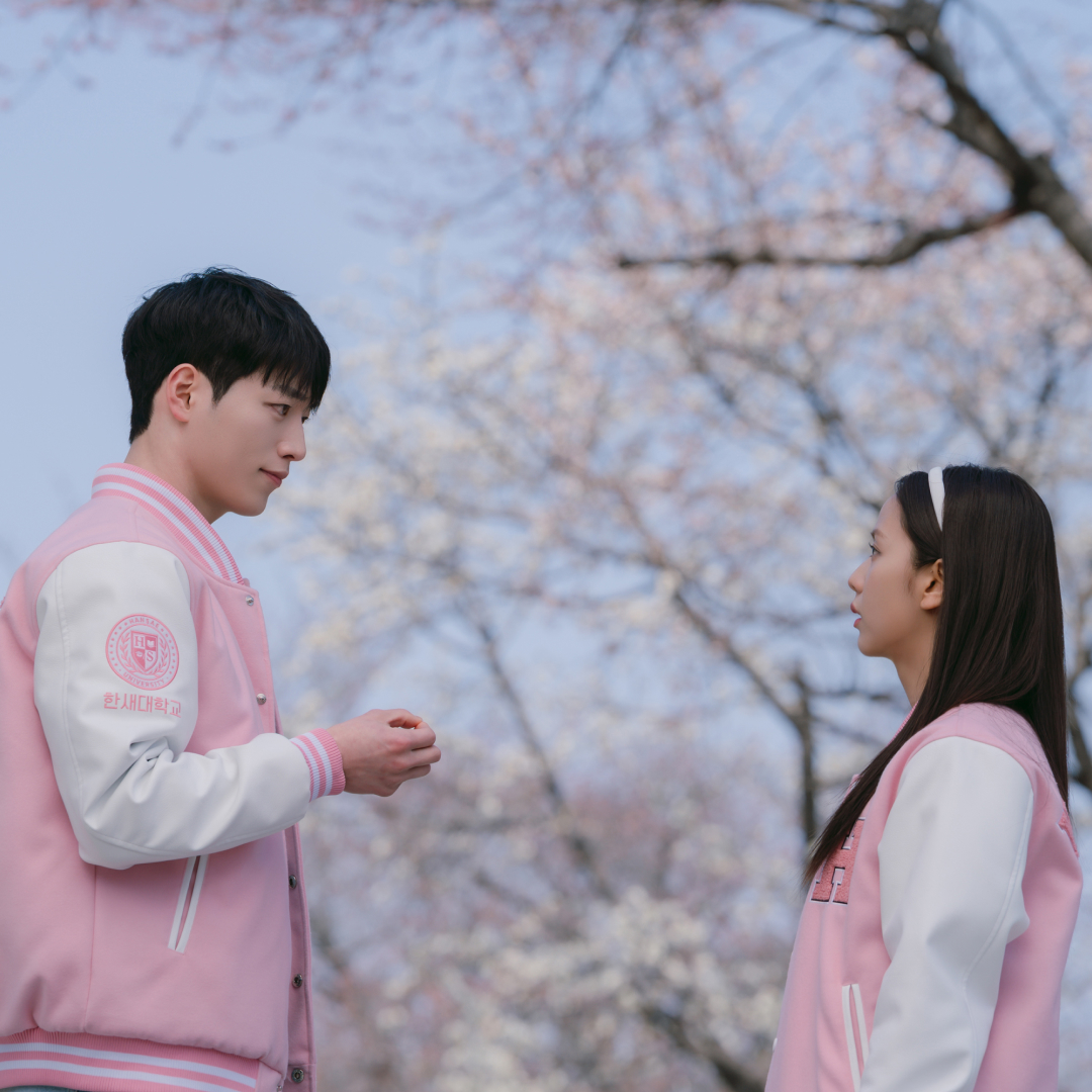 A man and a woman (Seo Kang-jun as Seo Eun-ho and Jisoo as Seo Mi-rae) wear pink and white Letterman jackets while standing outdoors in front of a cherry blossom tree, in 'Boyfriend on Demand.'