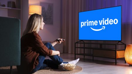A woman sits on the floor of a living room holding a remote up to a TV with the Prime Video logo on it. 