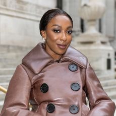 a black woman with dark hair pulled back into a bun posing in front of steps in a brown coat with black buttons
