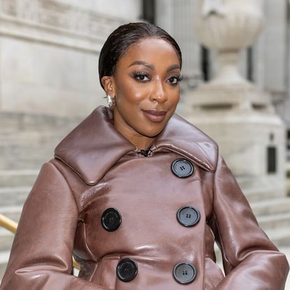 a black woman with dark hair pulled back into a bun posing in front of steps in a brown coat with black buttons
