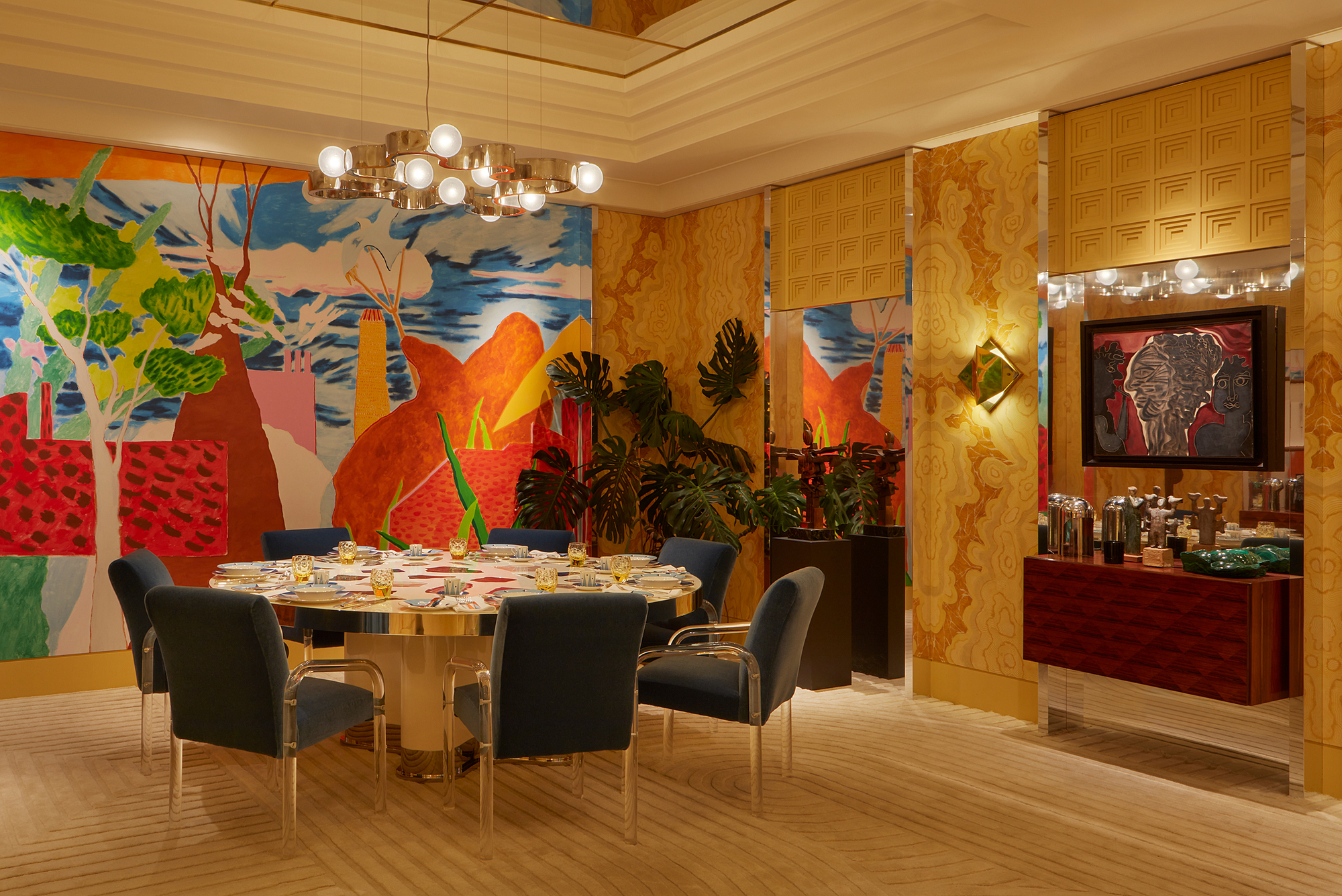 a yellow formal dining room with a round table and blue mohair chairs, yellow hand-painted wall panels and large mural, pendant light, stepped ceiling with mirrored panels, and drinks cabinet