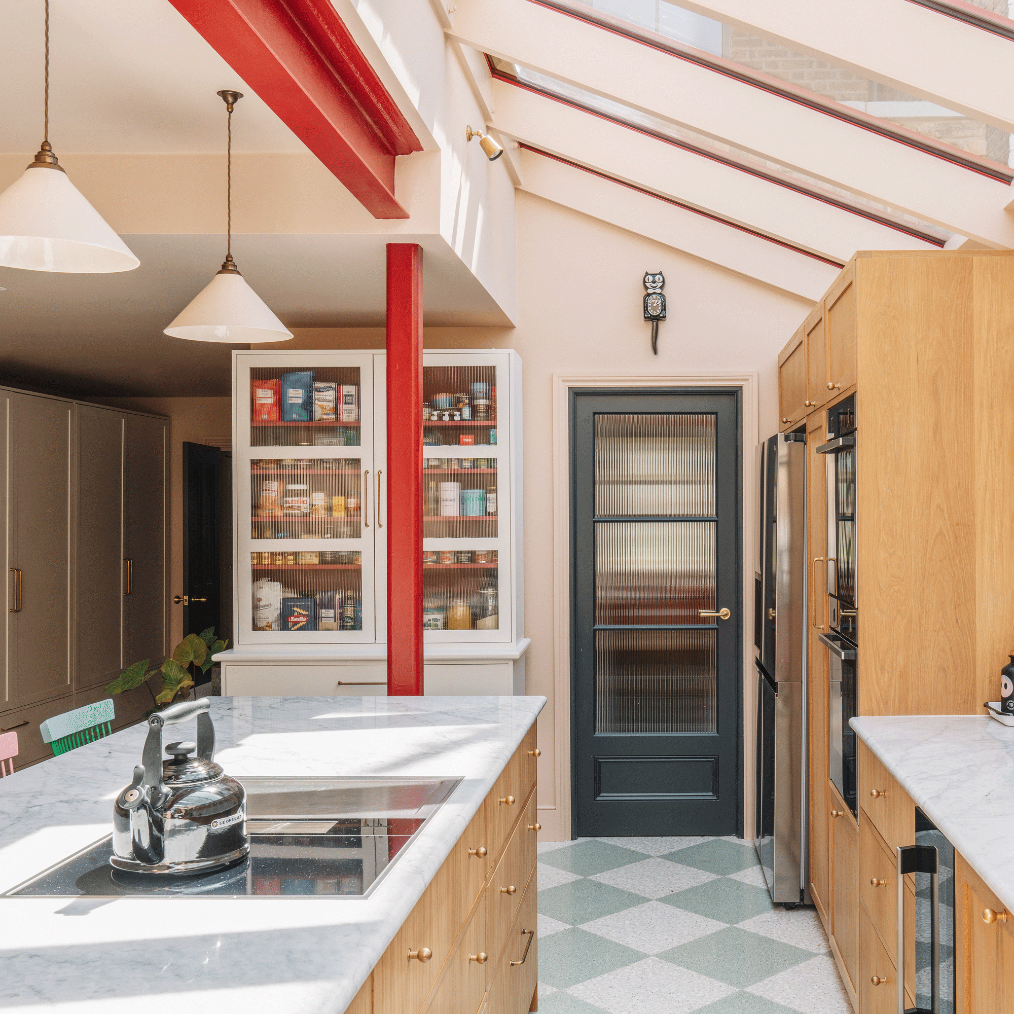 a kitchen with pale pink walls and wooden cabinets, a kitchen island with a marble countertop, pantry cupboard with reeded glass doors and a bright red pillar beside a black reeded glass door