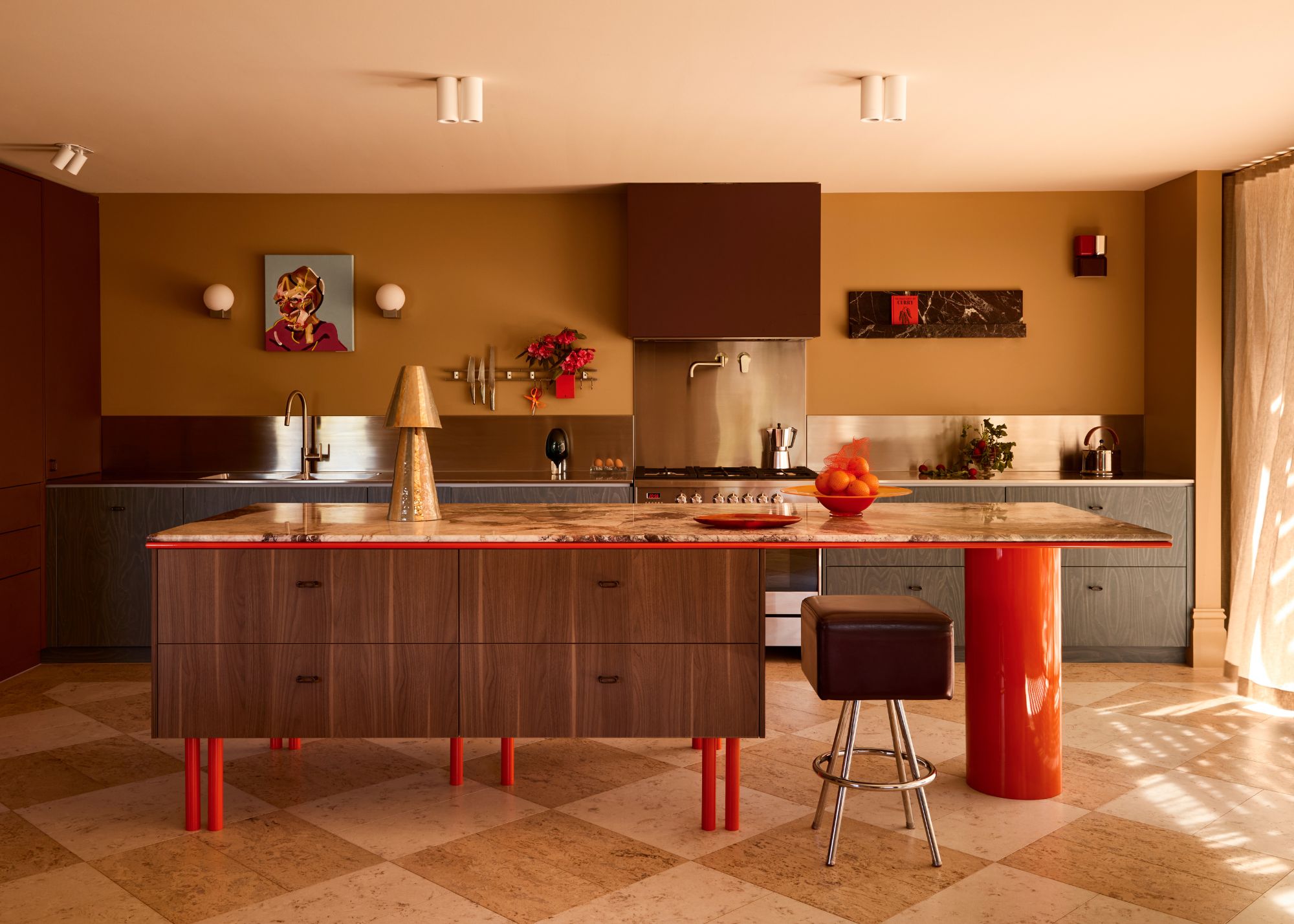 A modern kitchen with a kitchen island with vibrant lacquer red legs and a large circular leg on the seating side, with wood cabinetry and oxblood and blue cabinets against a mustard wall