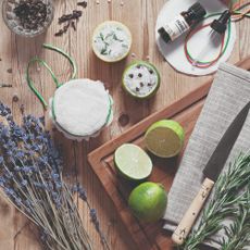 A wooden table with a wooden cutting board and halved limes, rosemary and lavender and a bottle of orange essential oil