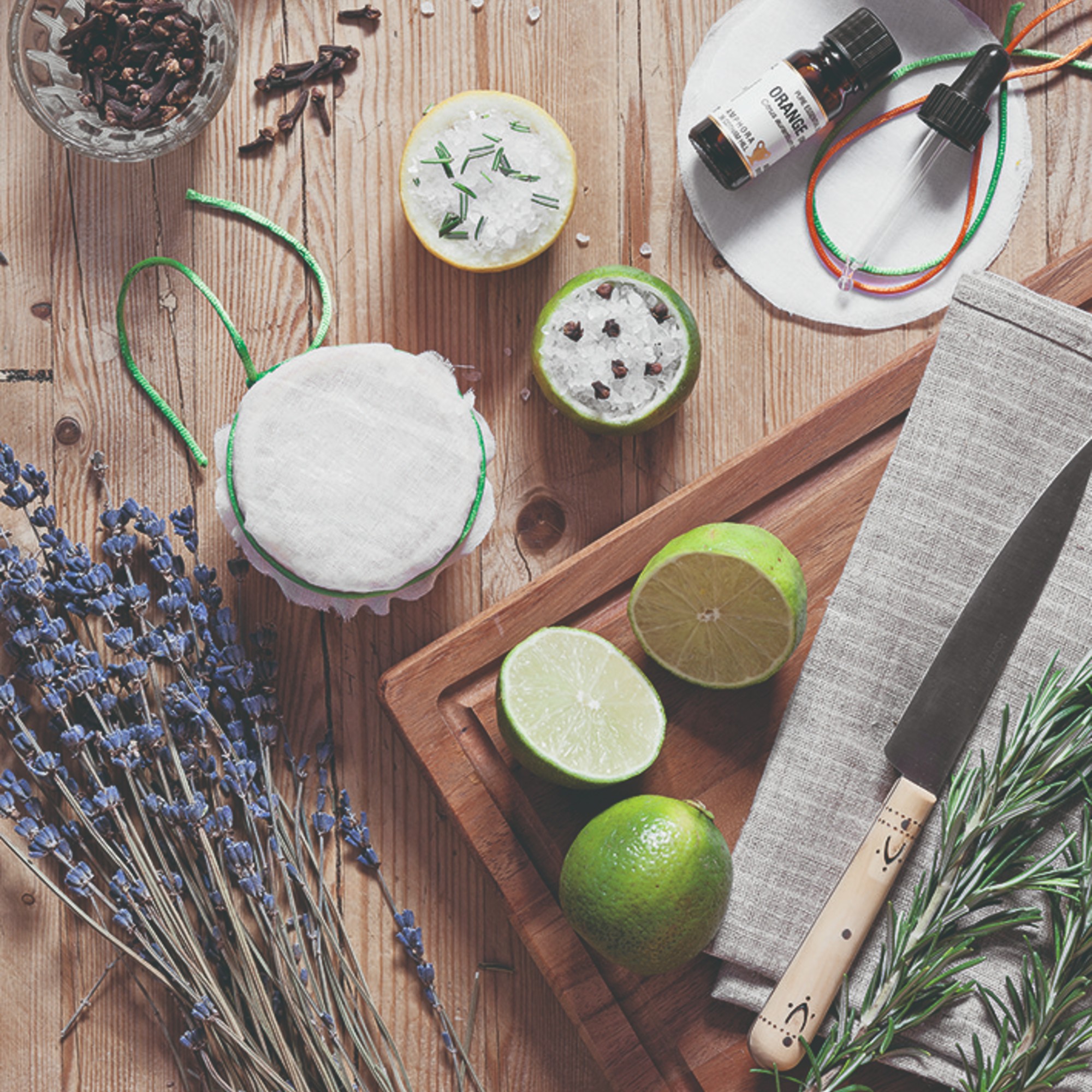 A wooden table with a wooden cutting board and halved limes, rosemary and lavender and a bottle of orange essential oil