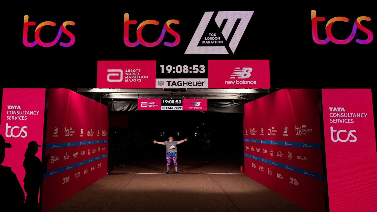 A London Marathon runner pauses under the gantry to celebrate finishing. It is nighttime. The time on the gantry reads 19:08:53